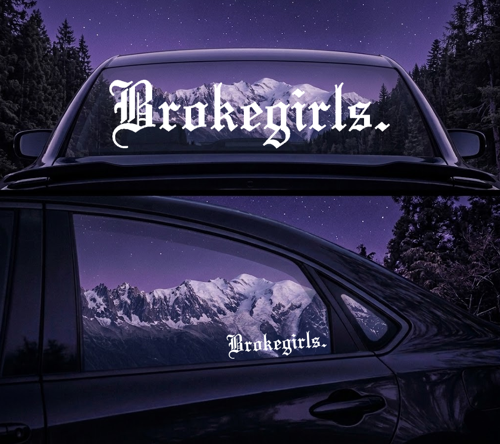 Car with 'Brokegirls' decal on the windshield and window, mountainous landscape in the background.
